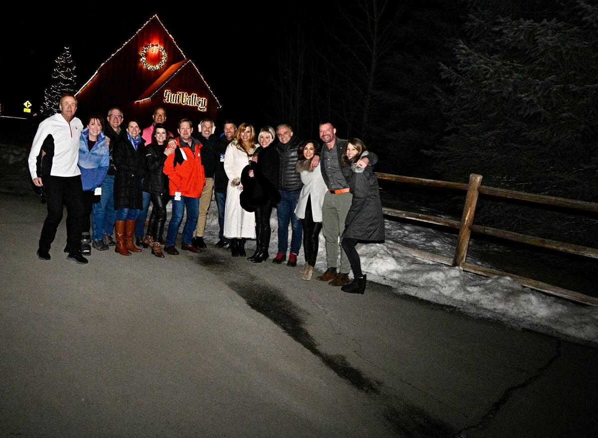A group in winter coats lined up along a snowy fence at night beneath the lit Sun Valley barn sign