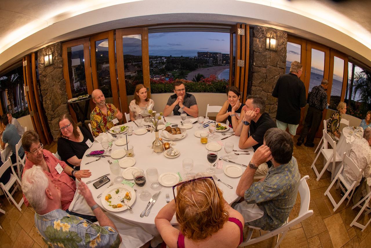 Round dinner tables in a room whose open windows look out over the coast at sunset