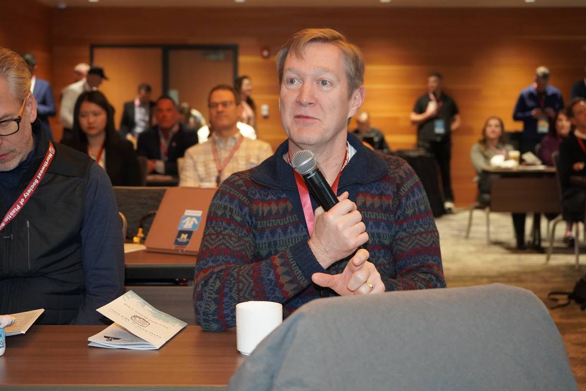 A seated attendee speaks into a handheld microphone during discussion, colleagues listening behind him