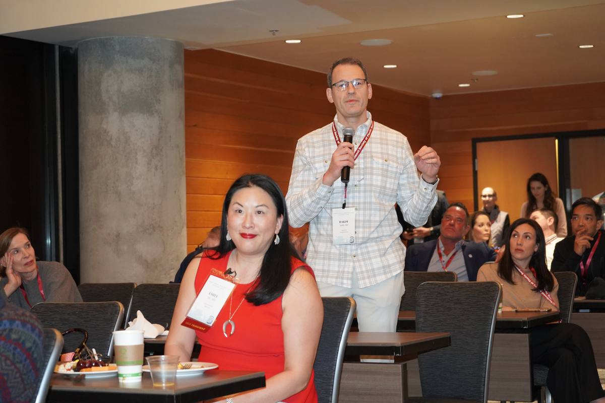 An attendee stands with a microphone to ask a question while others at the tables turn to listen