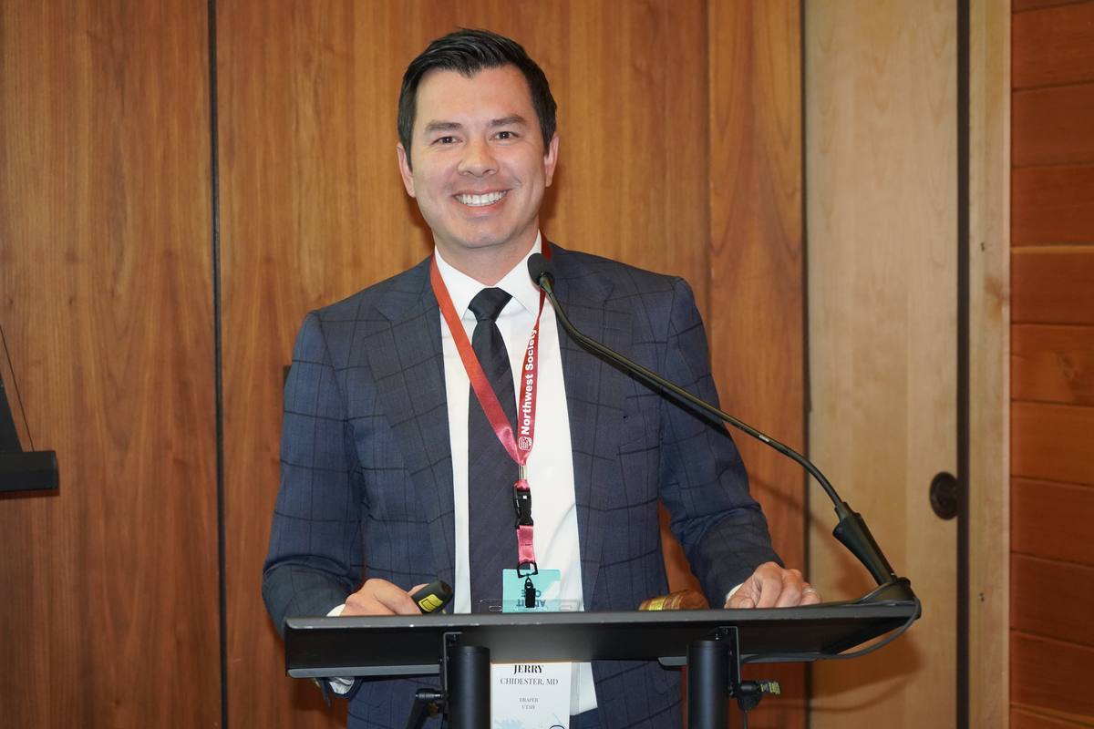 A smiling speaker at the lectern in the wood-panelled session room