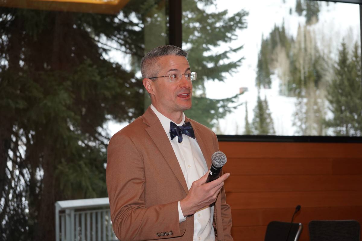 A speaker in a bow tie addresses the room with a microphone, snowy evergreens filling the window behind him
