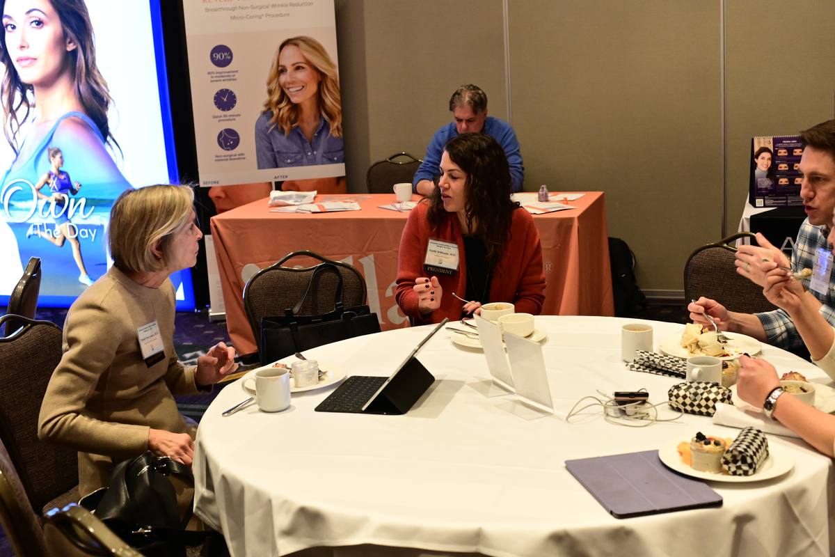 Two attendees in conversation over coffee at a table, an exhibitor’s display behind them