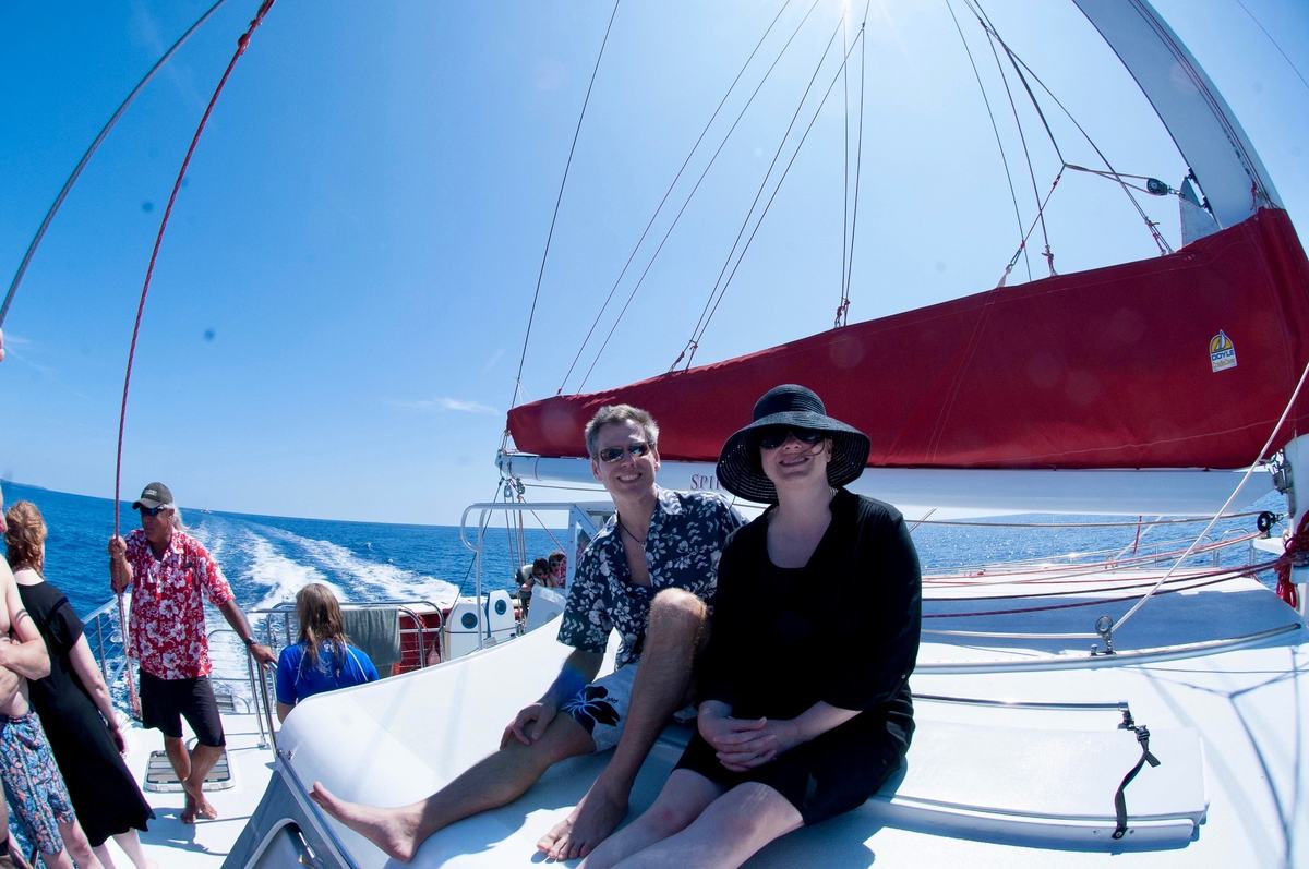 Two people relax on the bow of a sailing catamaran under a red sail, the wake bright behind them