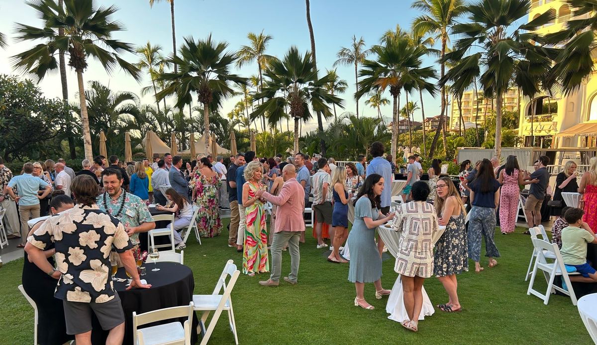 Members, partners, and children mingling at cocktail tables on a resort lawn under palm trees at sunset